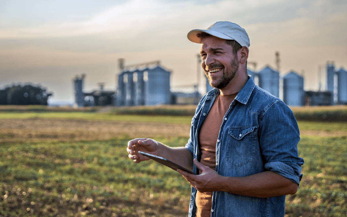 Farmer standing in a field holding a tablet, with grain bins in the background, representing agriculture and rural banking.